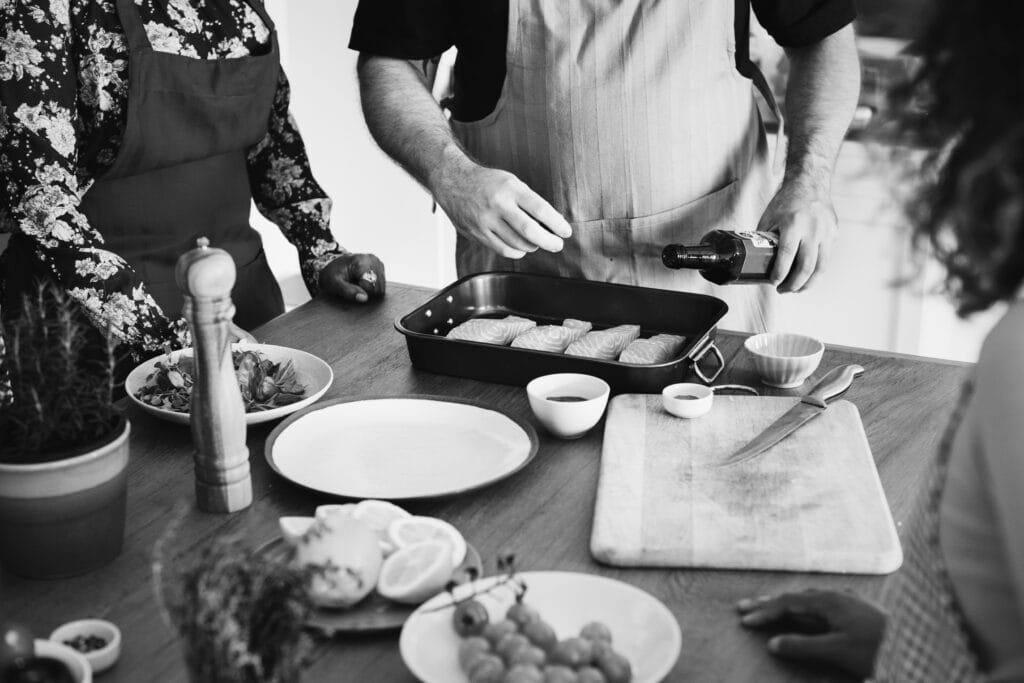Three people in aprons prepare a meal together, with one person pouring oil over food in a baking tray on a wooden counter—showcasing how shared spaces like the Campbell kitchen make cooking and kitchen cleaning enjoyable.