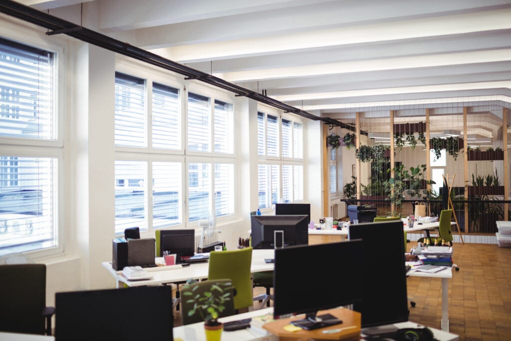 Modern open-plan office in Los Gatos with several desks, computers, and chairs, large windows letting in natural light, and green plants throughout the space—perfect for office managers seeking effective janitorial cleaning solutions.