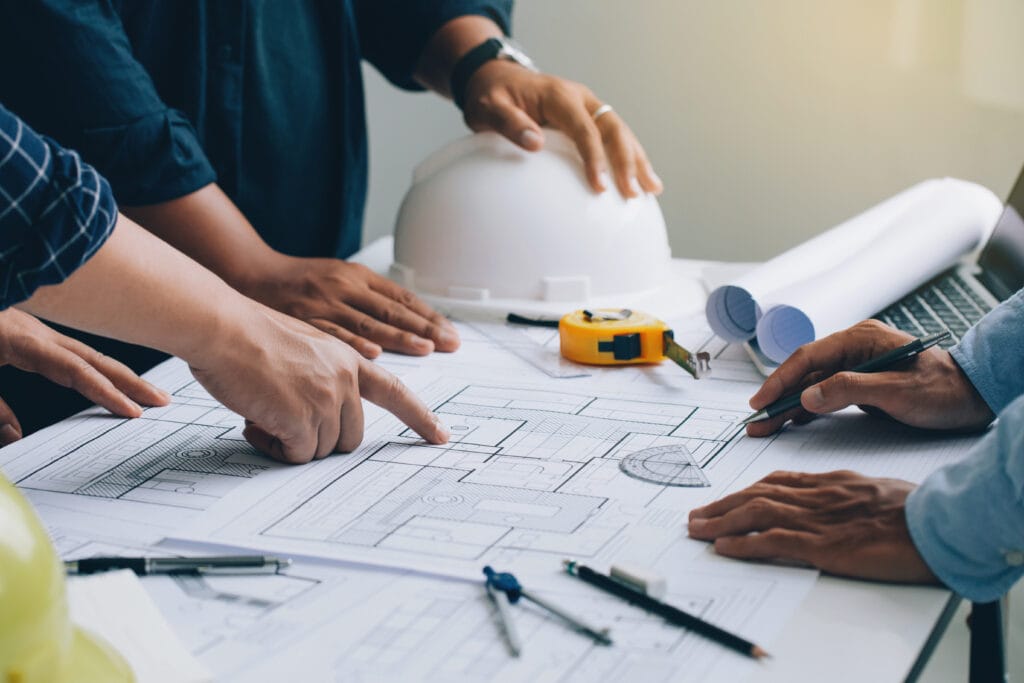 Several people review and discuss architectural blueprints on a table in Saratoga, surrounded by a hard hat, tape measure, and drafting tools—planning the next phase before post-construction cleaning begins.