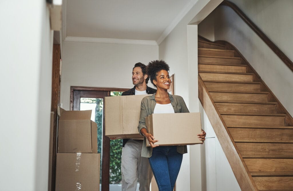 Two people carrying cardboard boxes walk through a hallway with a staircase and other moving boxes, suggesting they are moving into a new Saratoga home—perhaps with help from real estate agents or after a thorough roommate move-out cleaning.