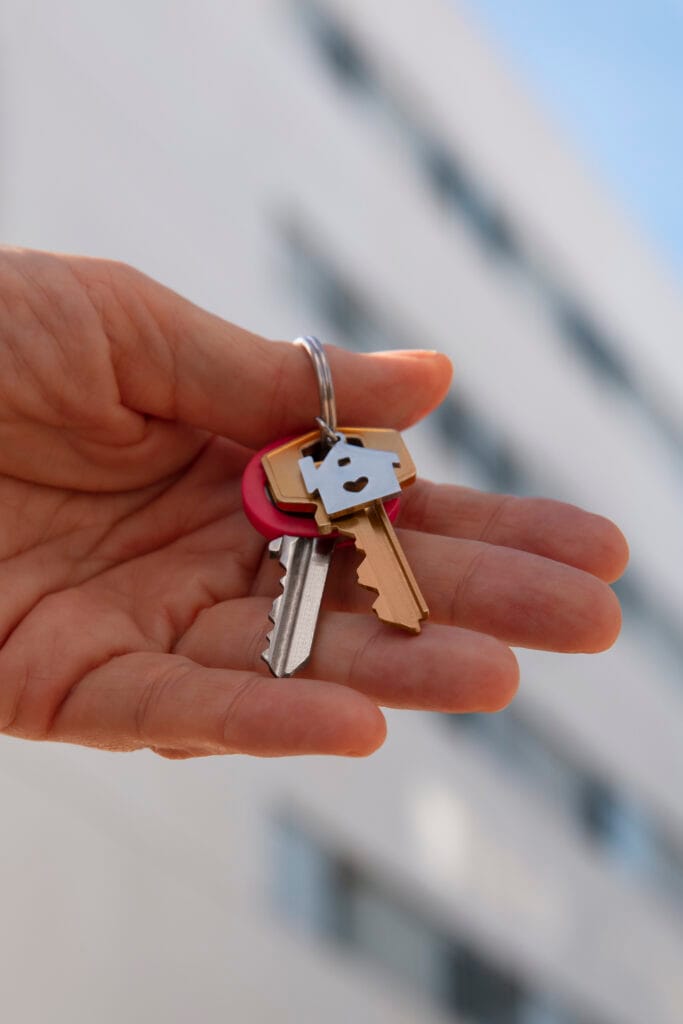 A hand holds a set of house keys on a keyring, symbolizing a spotless exit as real estate agents ensure every move-out cleaning in Cupertino leaves properties ready for new beginnings. A blurred building is visible in the background.