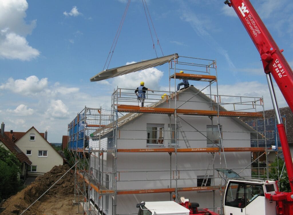 Builders in Santa Clara install a large roof panel onto a house using a crane, with scaffolding set up around the building under a partly cloudy sky as post-construction cleaning awaits.