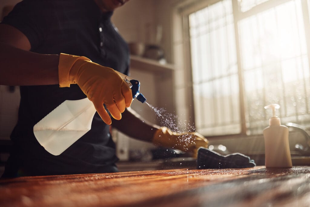 Person wearing yellow gloves sprays cleaning solution on a wooden surface, wiping it with a cloth in a sunlit kitchen. A soap dispenser is visible on the counter, reflecting the thoroughness of Los Gatos Deep Cleaning services for homeowners.