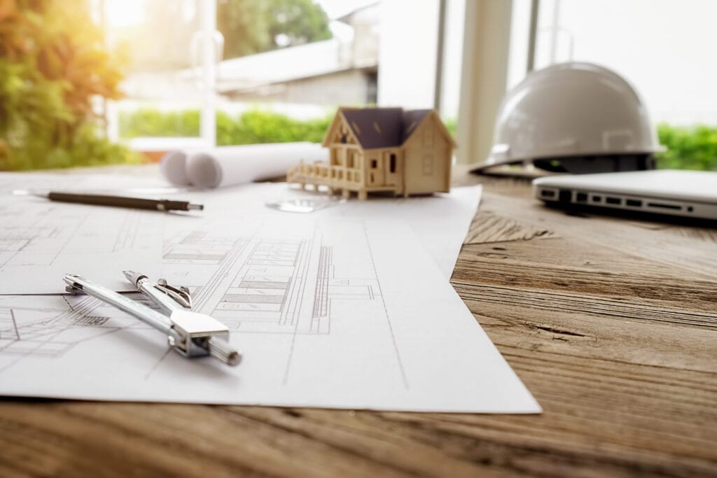 Architectural plans, compass, small house model, hard hat, and laptop on a wooden desk by a window in Saratoga, suggesting a business-ready construction or design workspace.