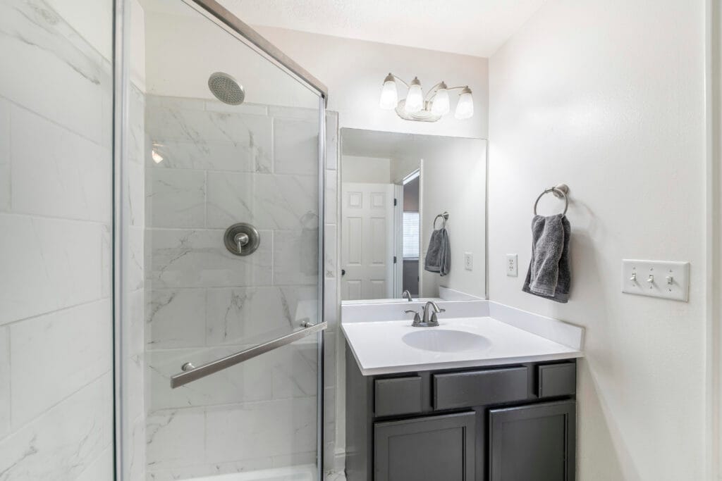 Modern bathroom in Cupertino featuring a glass-enclosed shower, white tile, single-sink vanity with dark cabinets, large mirror, and wall-mounted lighting—ideal for Property Managers seeking customized bathroom cleaning solutions.