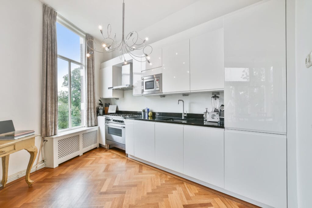 Modern kitchen with white cabinets, black countertop, stainless steel appliances, wooden herringbone floor, large window, and a contemporary chandelier—ideal for real estate agents staging homes in Saratoga.