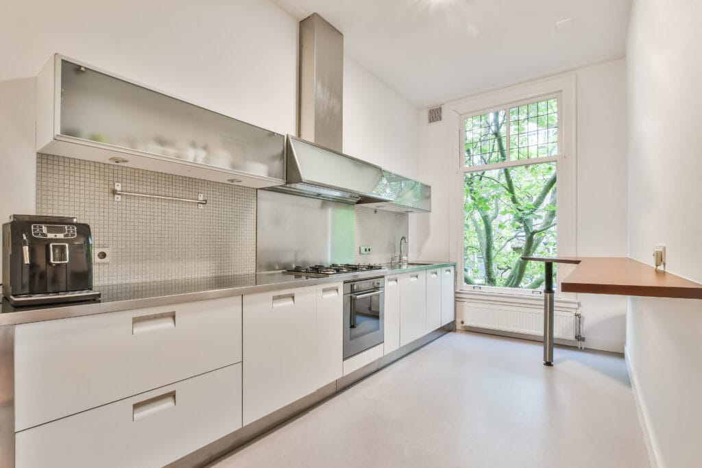 Modern kitchen with white cabinets, stainless steel countertops, built-in appliances, coffee machine, and a narrow bar table overlooking a leafy tree—perfect for Santa Clara homeowners seeking Kitchen-Only Cleaning solutions.
