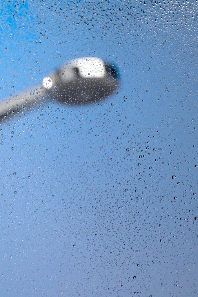 Close-up of a showerhead seen through wet, foggy glass with water droplets, set against a blue background—ideal inspiration for property managers focused on bathroom deep cleaning in Santa Clara County.
