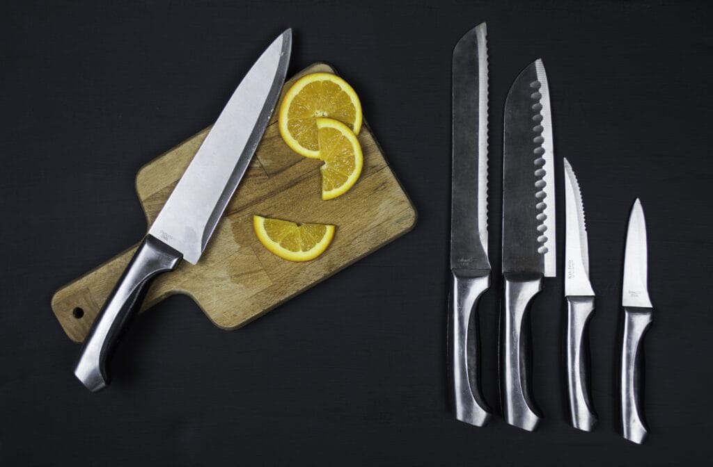 A wooden cutting board with a chef’s knife and orange slices on the left; four different knives arranged on a black surface to the right—ideal for shared spaces like Campbell, where kitchen cleaning is essential.