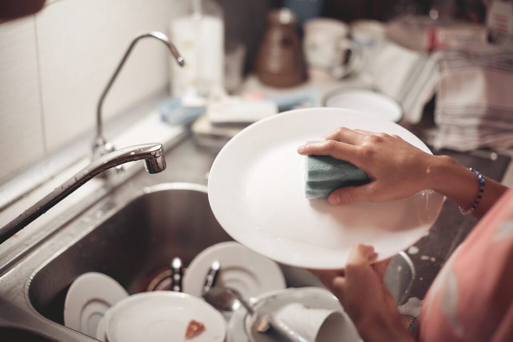 A homeowner washes a white plate with a green sponge at a kitchen sink filled with dirty dishes, focusing on kitchen-only cleaning.