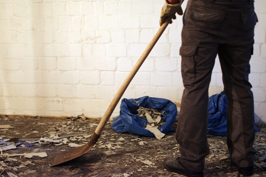 A homeowner in Saratoga, wearing gloves and work pants, uses a shovel to clean debris from the floor; blue bags filled with rubble show thorough post-construction cleaning against a white brick wall.