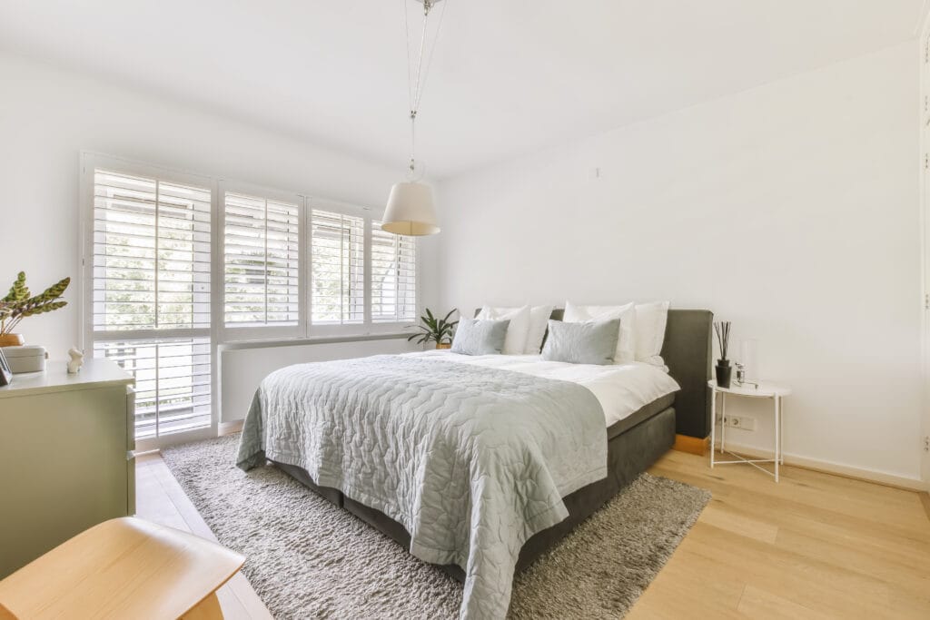Bright bedroom with a large bed, gray bedding, and indoor plants on light wood flooring. Sunlight streams through white shuttered windows—perfect after a Remodel Cleaning or Post-Renovation service in San Jose.