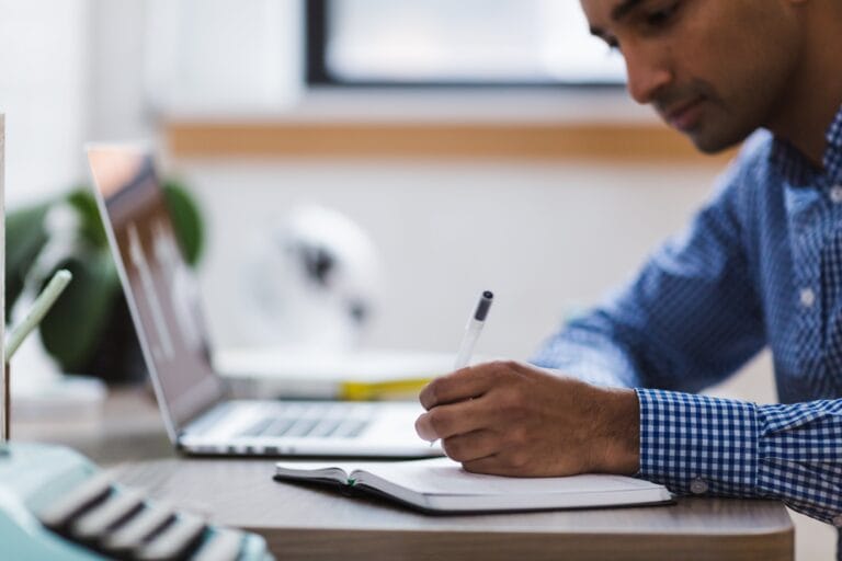 A person in a blue checkered shirt writes in a notebook while working at a desk with an open laptop, perhaps planning Kitchen-Only Cleaning services for Saratoga property managers.