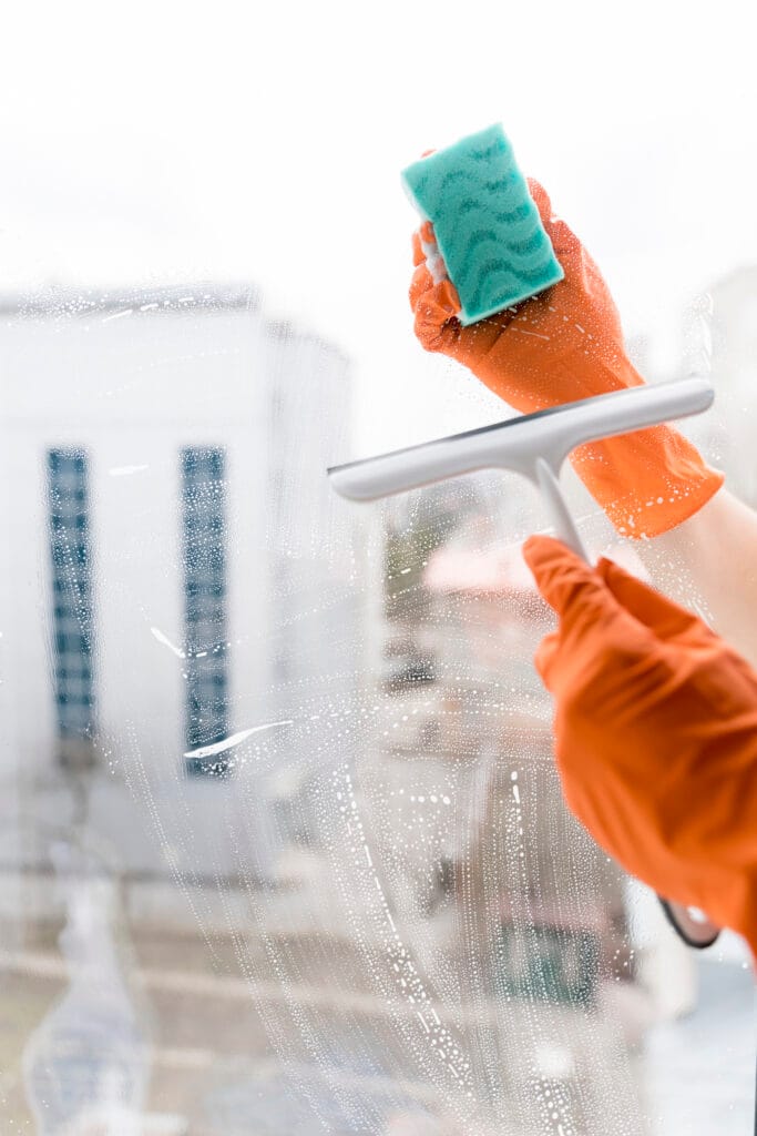 A person wearing orange gloves cleans a glass window with a sponge and squeegee as part of a Partial Renovation Cleaning, with buildings visible in the blurry background—perfect for Campbell homeowners.