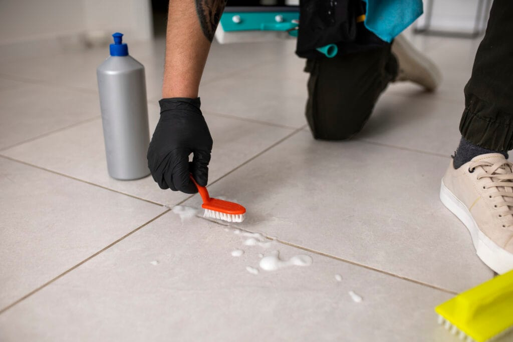 Person wearing a black glove scrubs a tiled floor with a brush and cleaning solution; ideal for bathroom deep cleaning in Santa Clara—essential for real estate agents preparing homes. A bottle of cleaner and supplies are visible nearby.