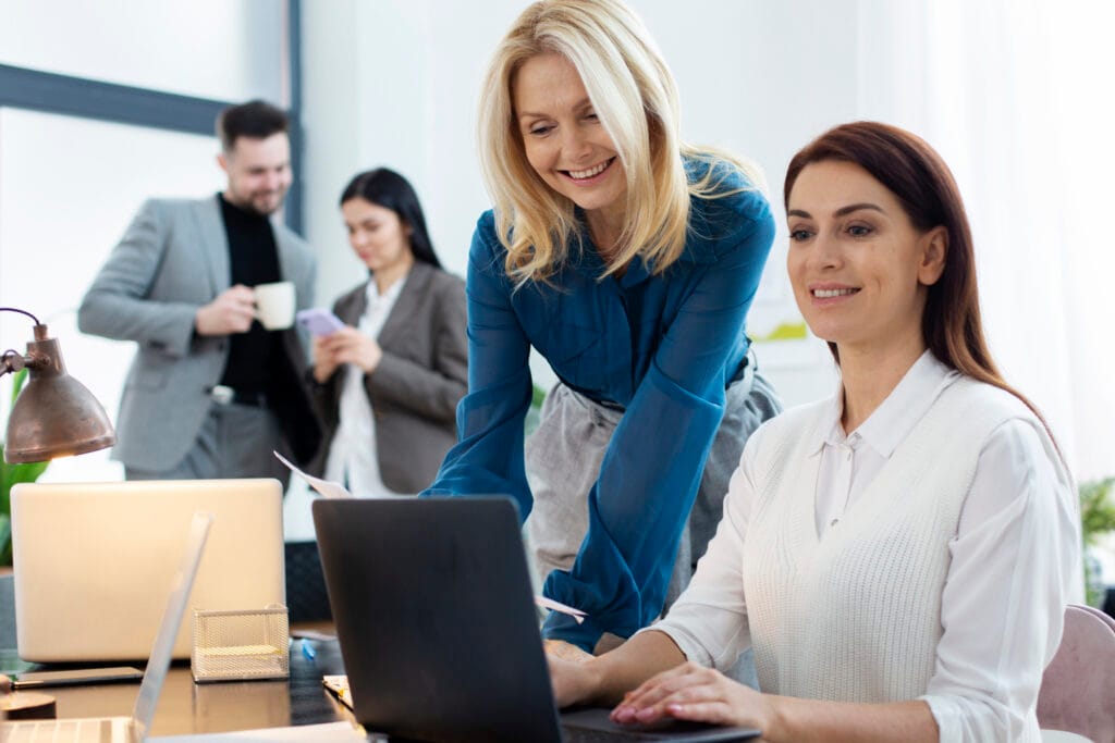 Two women work together at a laptop in a Cupertino office, while a man and another woman talk and hold drinks in the background—ideal teamwork for Office Managers after Post-Construction Cleaning.