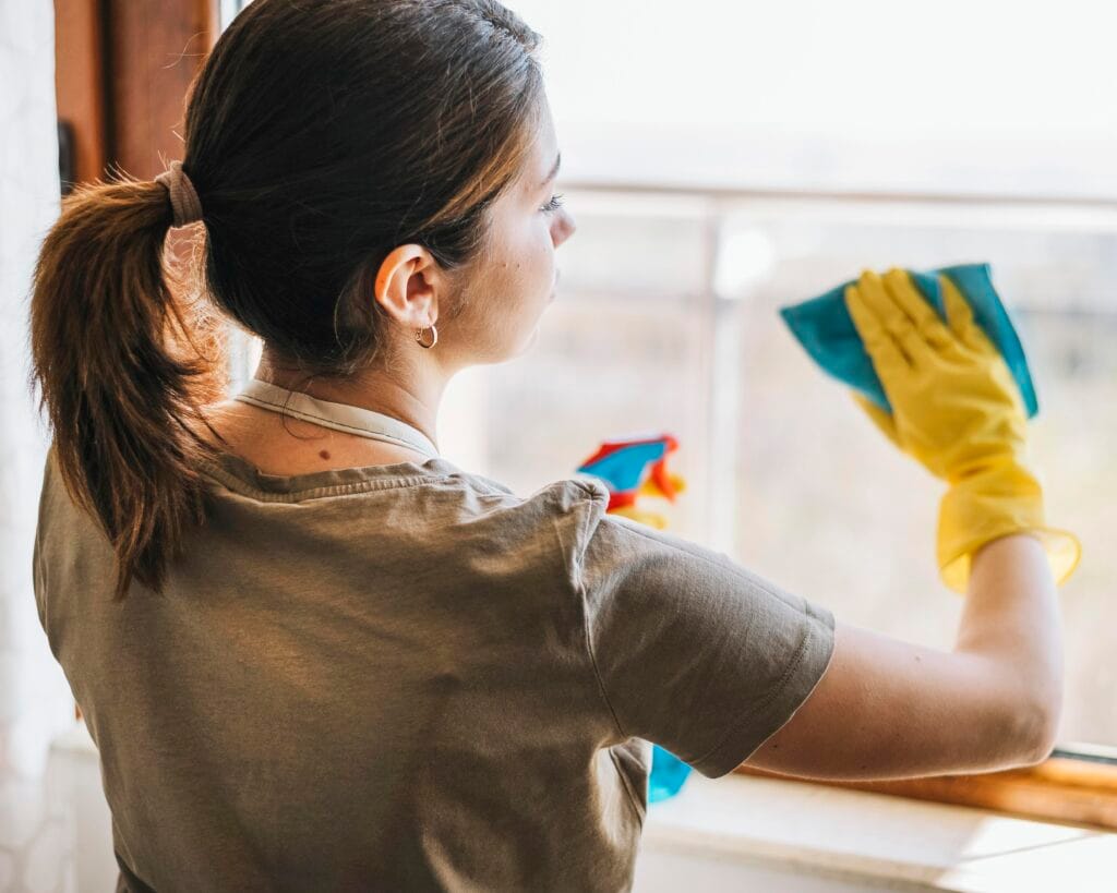 A woman wearing yellow gloves cleans a window with a blue cloth and spray bottle during the day, demonstrating how Cupertino Deep Cleaning Services can help property managers restore your space to pristine condition.