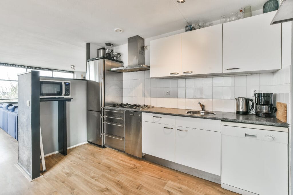 Modern Santa Clara kitchen with white cabinets, stainless steel appliances, a microwave on a stand, coffee maker, and kettle on a black countertop with wood flooring—ideal for homeowners seeking Kitchen-Only Cleaning.