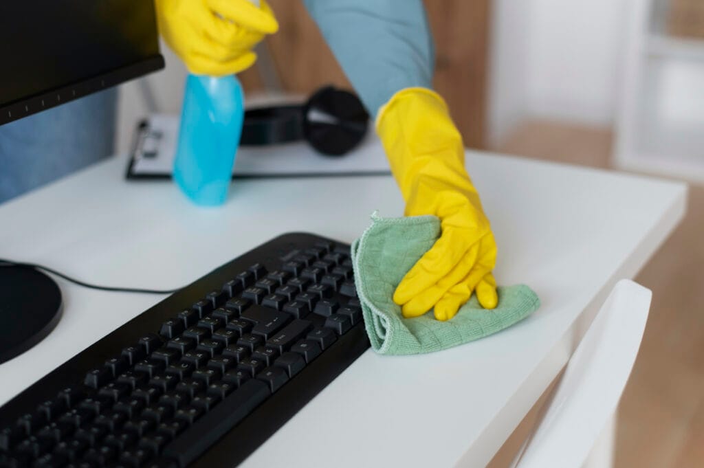 Wearing yellow gloves, a person provides expert office care by cleaning a desk near a keyboard with a green cloth and blue spray bottle.