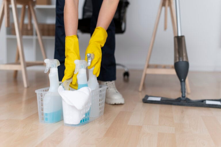 A person in yellow gloves places cleaning supplies in a basket on a wooden floor, ready for post-construction cleaning—ideal for Santa Clara office managers seeking spotless workspaces. A mop stands nearby, completing the scene.