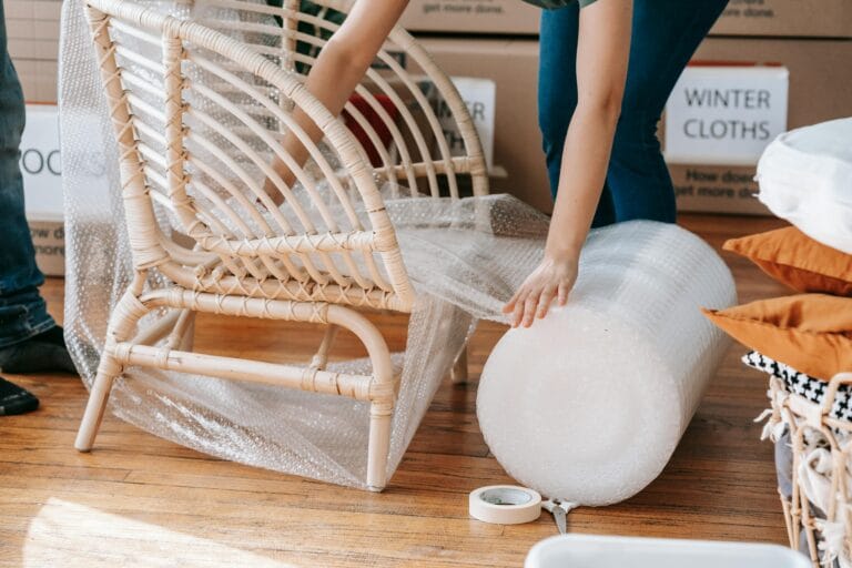 A person wraps a wicker chair in bubble wrap with tape and scissors on the wooden floor, surrounded by moving boxes—ensuring a Spotless Exit for their Saratoga home.