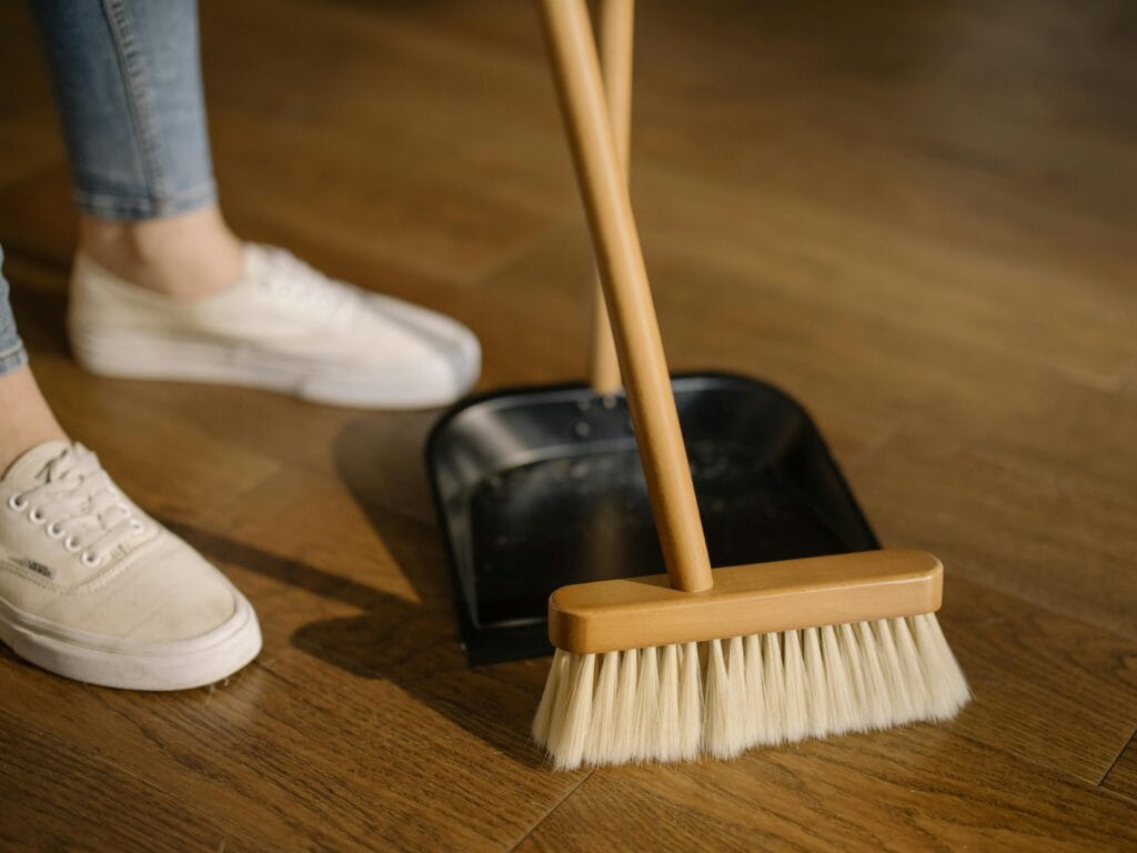 A person wearing white sneakers sweeps dirt into a black dustpan with a broom on a wooden floor during partial renovation cleaning in Santa Clara.