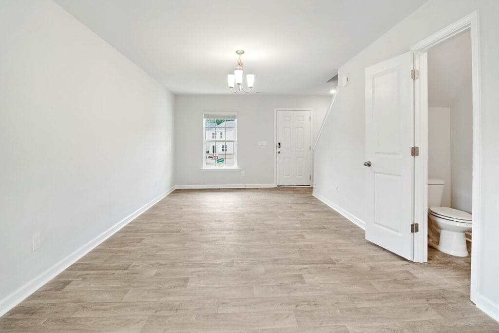Bright empty room in San Jose features light wood flooring, white walls, a ceiling light fixture, window, front door, staircase, and an open door to a small bathroom—ideal for real estate agents or after a roommate move-out cleaning.