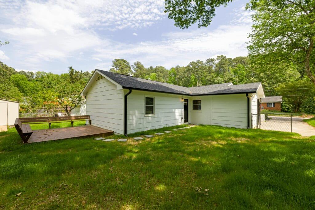 A single-story white house with black trim, a small wooden deck, and a grassy backyard bordered by trees under a partly cloudy sky—builders have ensured it’s jobsite ready for comfortable living.