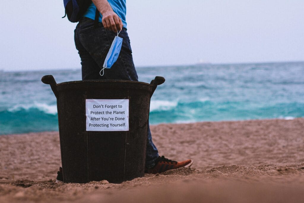 A person stands on a beach in Saratoga holding a face mask near a trash bin with a sign that reads, "Don't forget to protect the planet after you're done protecting yourself. Embrace a spotless exit—keep beaches clean!.