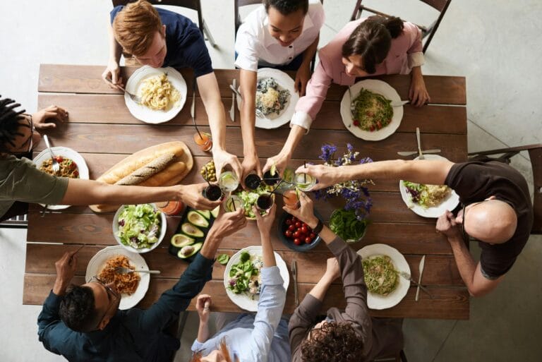 Eight people sit around a wooden table in Saratoga, plates of food and salads before them, raising their glasses together in a toast after enjoying the comfort of a kitchen-only cleaning—perfect for property managers seeking spotless spaces.