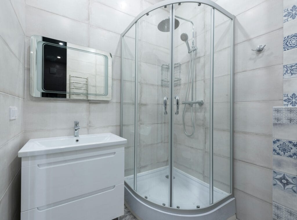 Modern bathroom with a glass-enclosed corner shower, white vanity with sink, wall-mounted mirror, and light-colored tiled walls—ideal for Santa Clara real estate agents seeking homes ready after a professional bathroom deep cleaning.