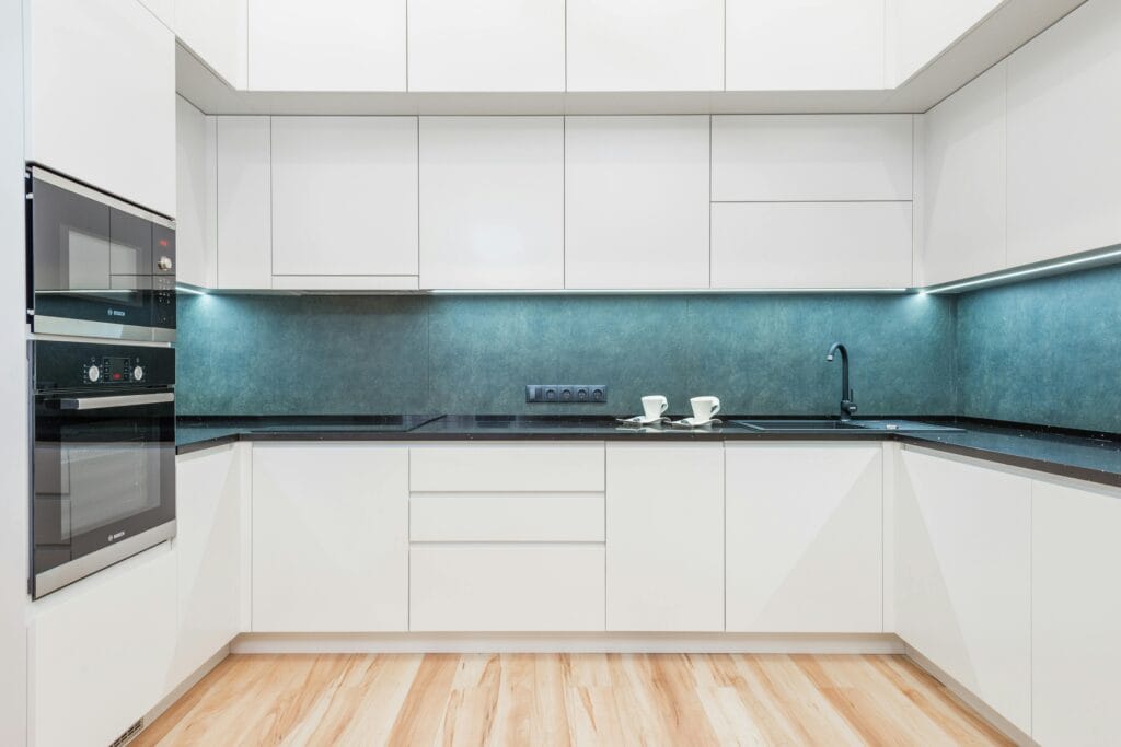 Modern kitchen in San Jose with white cabinets, black countertops, built-in oven, sink, and two cups on the counter, featuring wood flooring and under-cabinet lighting—ideal for real estate agents or roommate move-out cleaning.