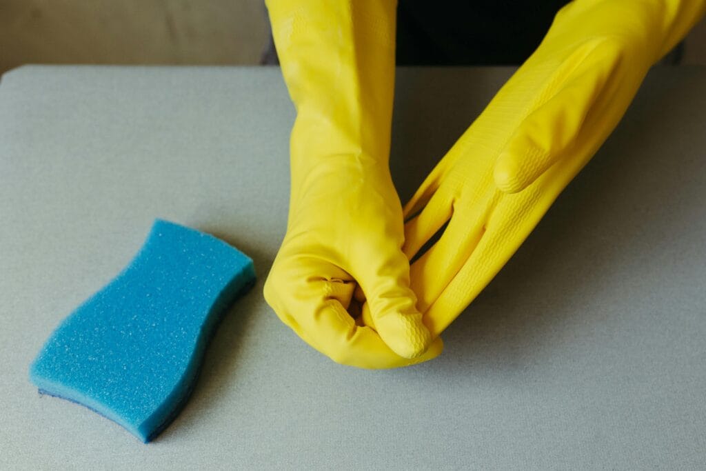 Person putting on yellow rubber gloves next to a blue cleaning sponge on a gray surface, ready for some serious home cleaning.