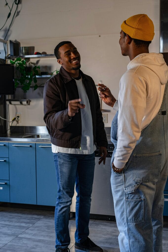 Two men stand in a kitchen, facing each other and smiling while having a conversation. One wears a brown jacket, the other sports a yellow beanie and denim overalls—typical homeowners in Santa Clara County enjoying their space.