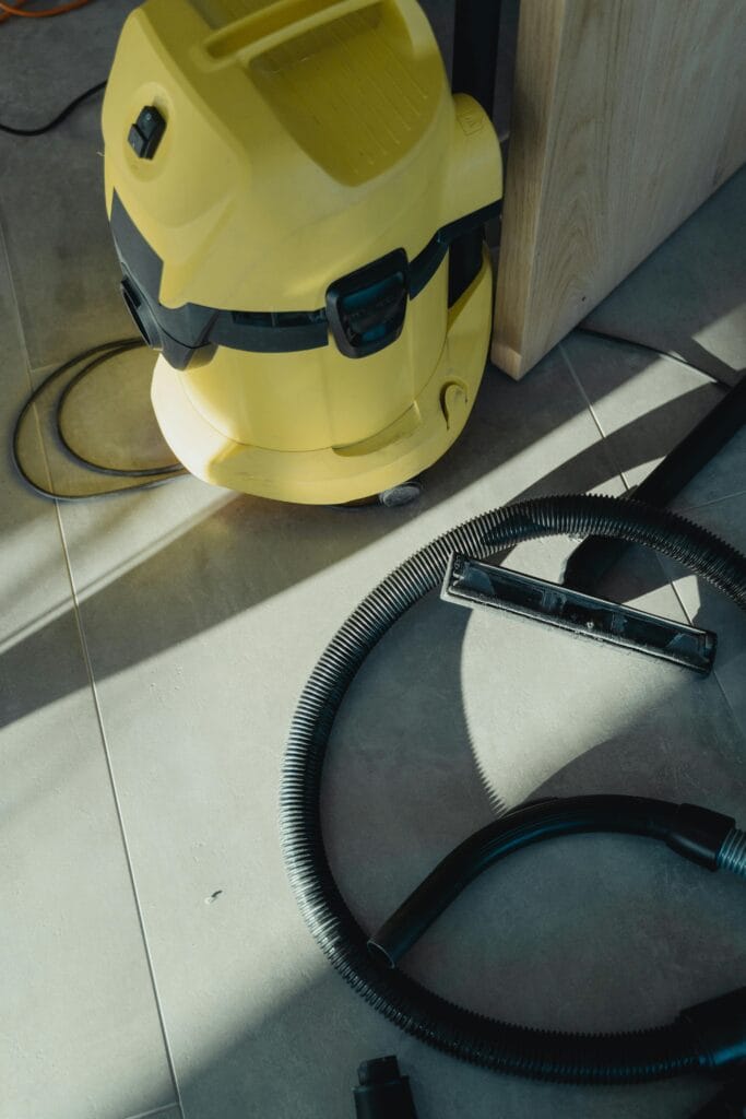 A yellow wet and dry vacuum cleaner with a detached hose and floor nozzle sits on a tiled floor in sunlight, ready for post-construction cleaning in Santa Clara County.