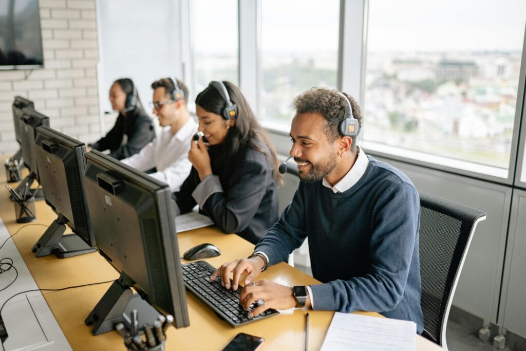 Four customer service representatives wearing headsets work at computers in a modern San Jose office with large windows, benefiting from professional janitorial cleaning that ensures a spotless and inviting workspace.