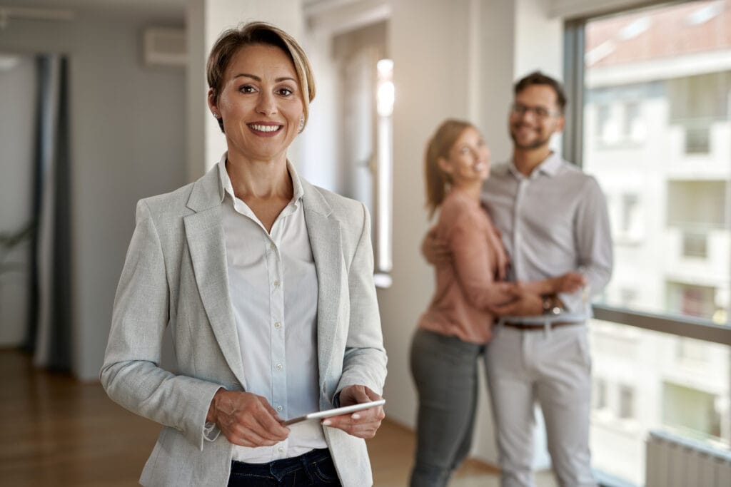 A real estate agent stands in the foreground holding a tablet, while a smiling couple embraces in the background inside a modern apartment—an ideal space for property managers seeking Cupertino Deep Cleaning Services.
