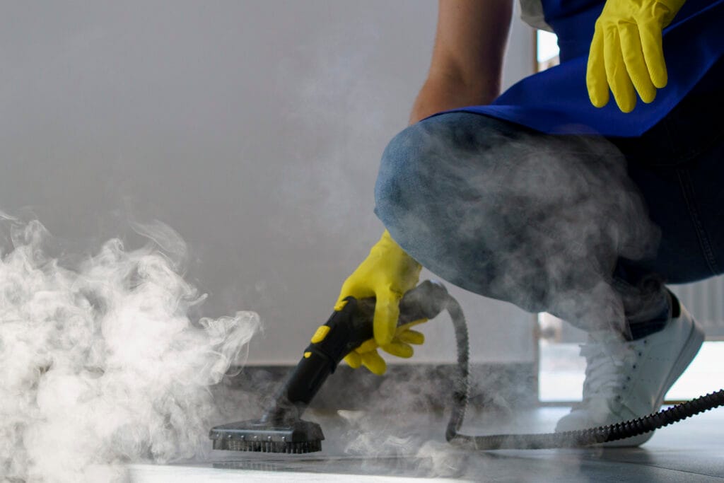 Wearing yellow gloves, a person uses a handheld steam cleaner on a tiled floor, with visible steam rising during post-renovation cleaning in Santa Clara.
