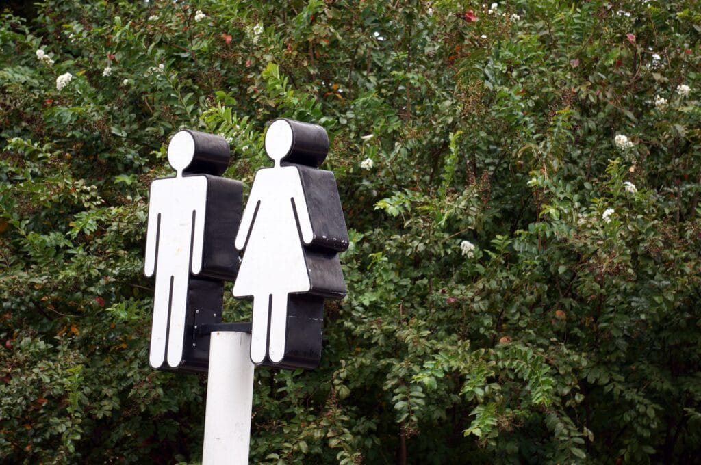 A restroom sign with male and female symbols is mounted on a post in front of dense green foliage, marking facilities commonly needed by builders and crews during jobsite cleaning.