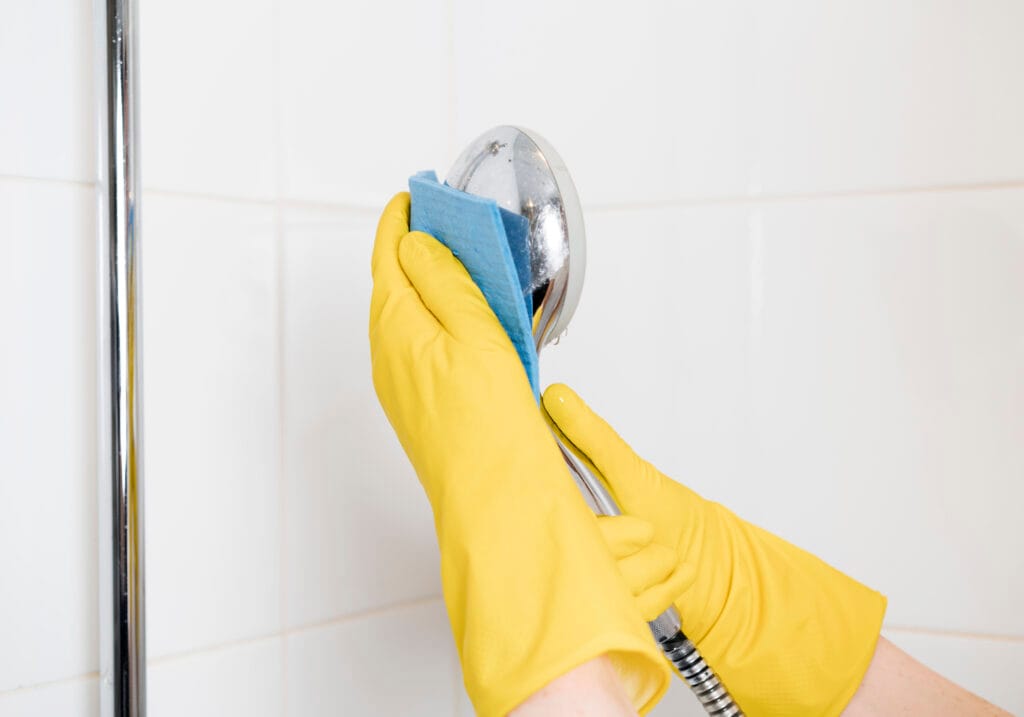 Person wearing yellow rubber gloves cleaning a showerhead with a blue cloth against a white tiled background, perfect for showcasing bathroom deep cleaning standards real estate agents in Santa Clara look for.