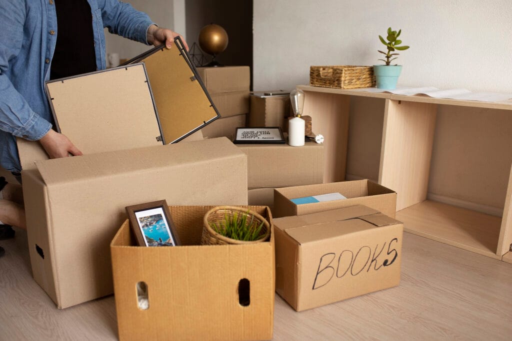 A person packs cardboard boxes with books, a framed photo, and a potted plant—empty shelf and various household items in the background—capturing the moving day hustle common in San Jose, especially after a roommate move-out cleaning.