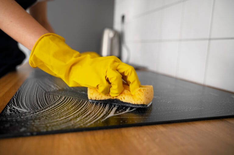 A person wearing a yellow glove cleans a stovetop with a sponge, creating soapy streaks on the surface—a glimpse into the meticulous results homeowners can expect from Los Gatos Deep Cleaning.