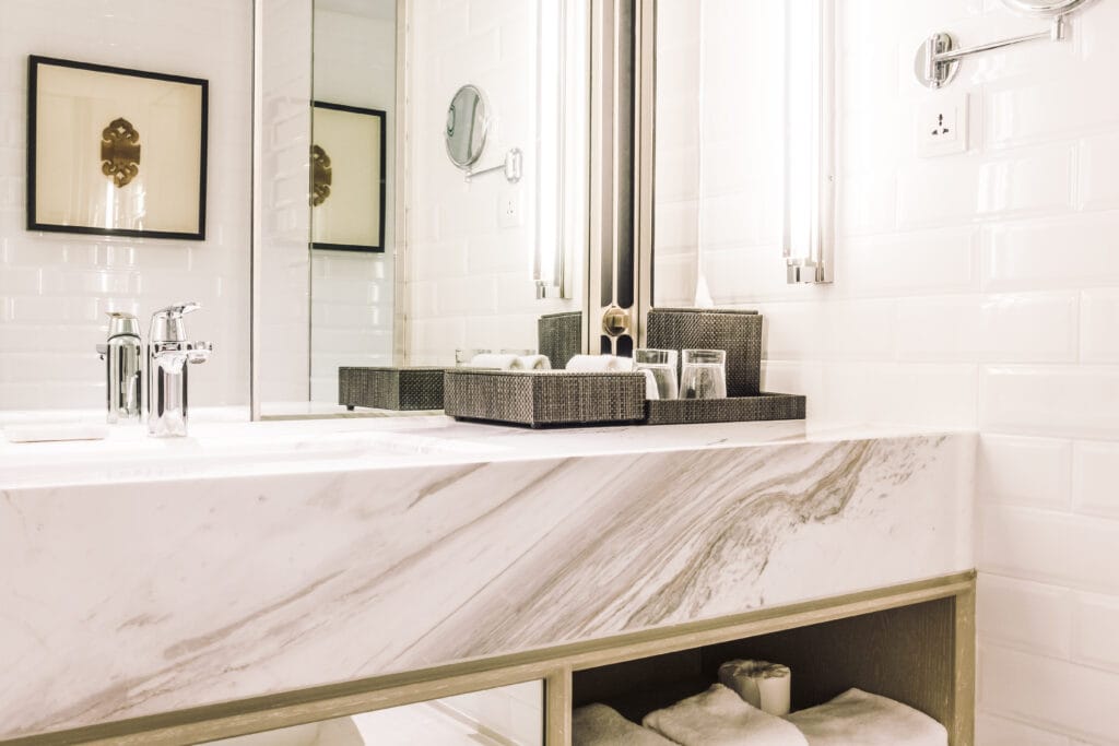 Modern bathroom with a marble countertop, organized tray with toiletries and glasses, towels on a shelf below, and white tiled walls—perfect for showcasing after Customized Bathroom Cleaning in Saratoga.
