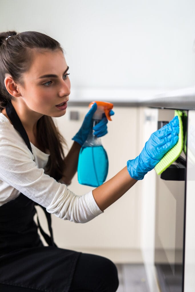 A woman wearing blue gloves uses a spray bottle and cloth to clean a kitchen appliance, showcasing the attention to detail offered by Cupertino Deep Cleaning Services—an ideal solution for busy property managers.