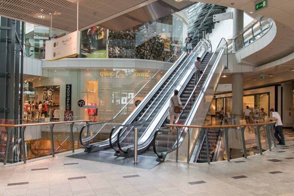 A shopping mall interior with two escalators going up, a few people using them, and various retail stores visible in the background, appears jobsite ready—builders have ensured everything is spotless with final cleaning San Jose professionals.