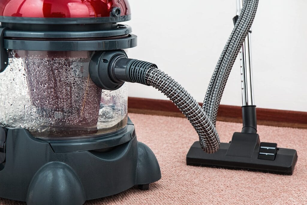 A vacuum cleaner with a transparent water tank and attached hose is positioned on pink carpet next to a wall and baseboard, ready for post-construction cleaning in a San Jose commercial setting.