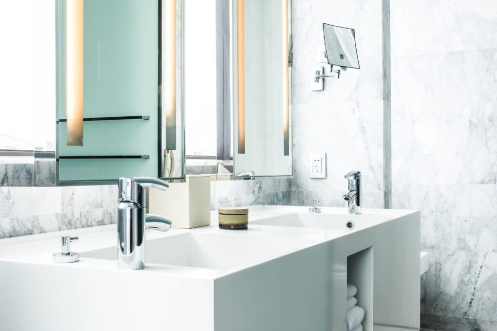 Modern white bathroom with a double sink, chrome faucets, a large mirror, wall-mounted lights, and neatly stacked towels on an open shelf—perfectly maintained with professional Bathroom Cleaning Saratoga services.