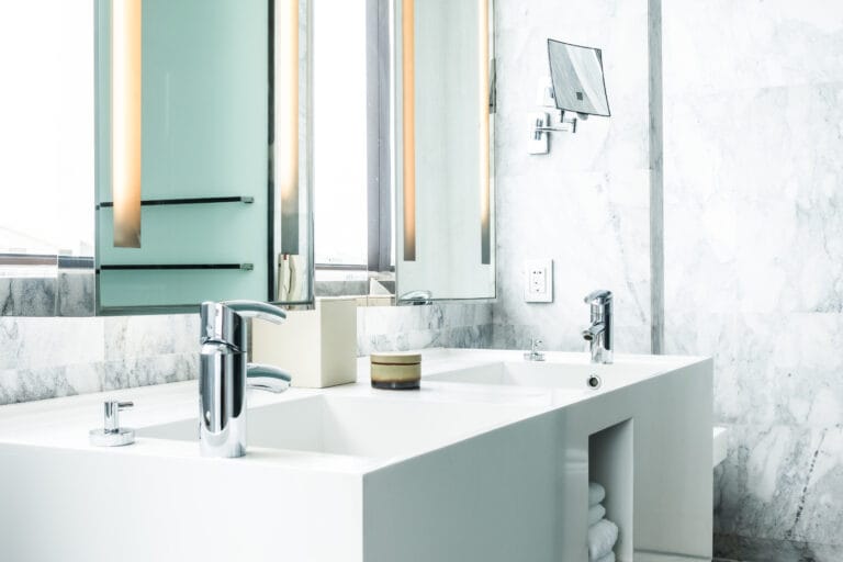Modern white bathroom with a double sink, chrome faucets, a large mirror, wall-mounted lights, and neatly stacked towels on an open shelf—perfectly maintained with professional Bathroom Cleaning Saratoga services.