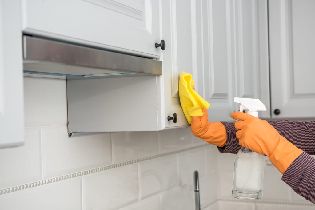 A person wearing orange gloves cleans a white kitchen cabinet with a yellow cloth and spray bottle—perfect for Move-Out Cleaning in Los Gatos. White tiled backsplash and cabinets are visible, making it ideal for Property Managers seeking spotless results.