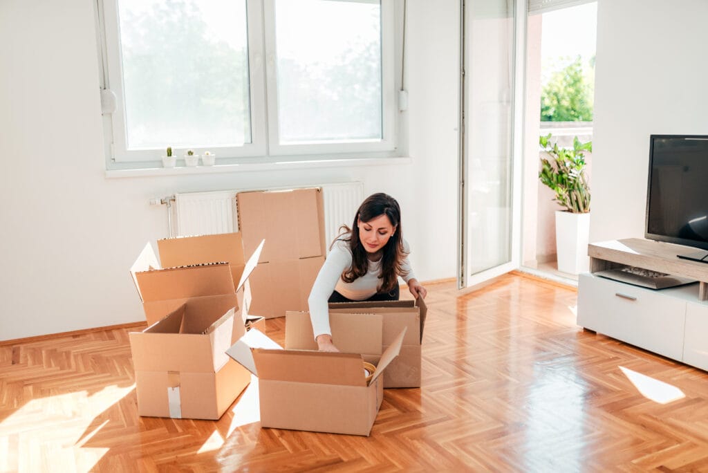 A woman sits on the floor of a bright Saratoga room, packing items into cardboard boxes, with several open boxes around her—preparing for a roommate move-out cleaning.